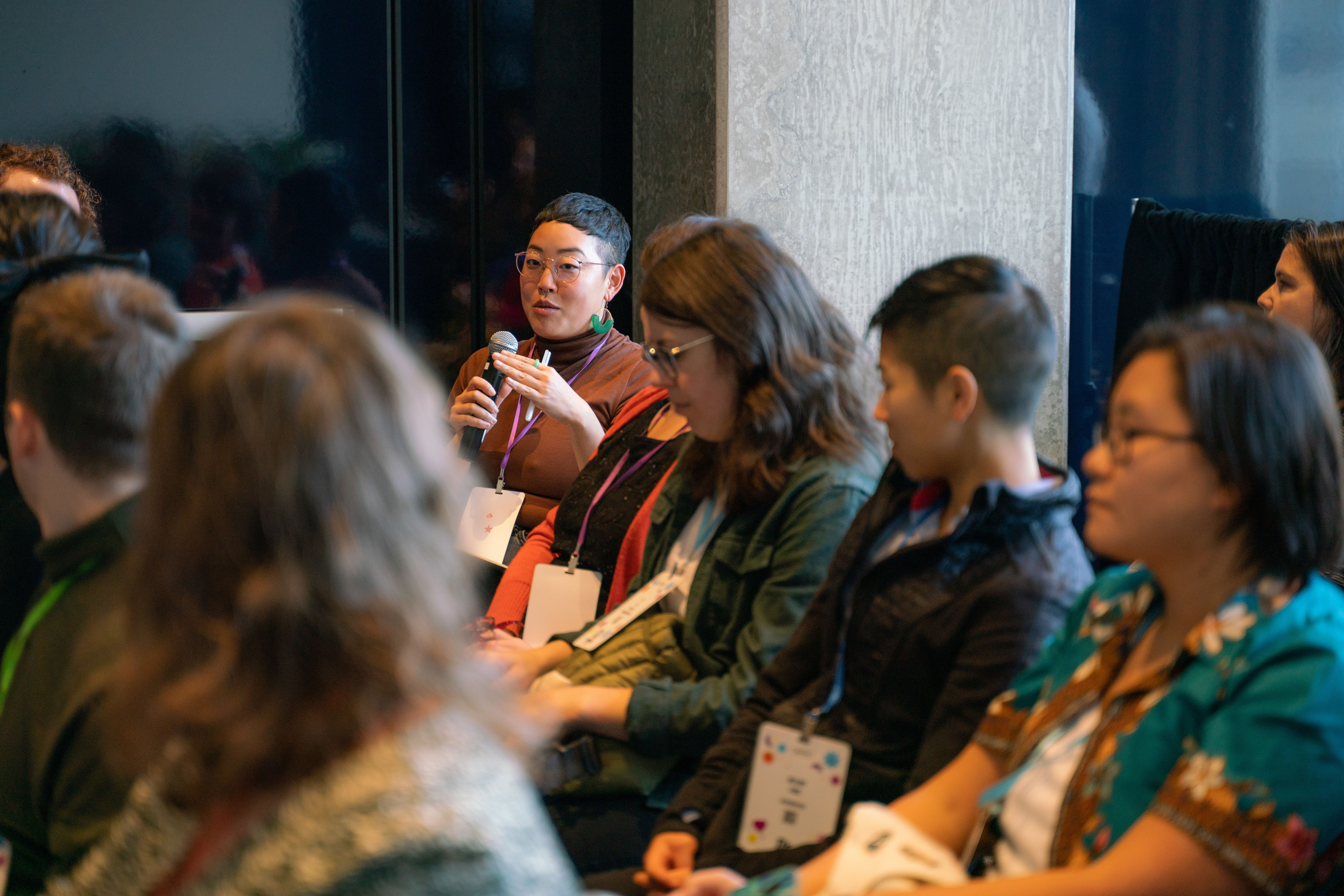 A diverse group sits in a discussion circle. One person speaks into a microphone, engaging an attentive audience. The atmosphere is focused and collaborative.