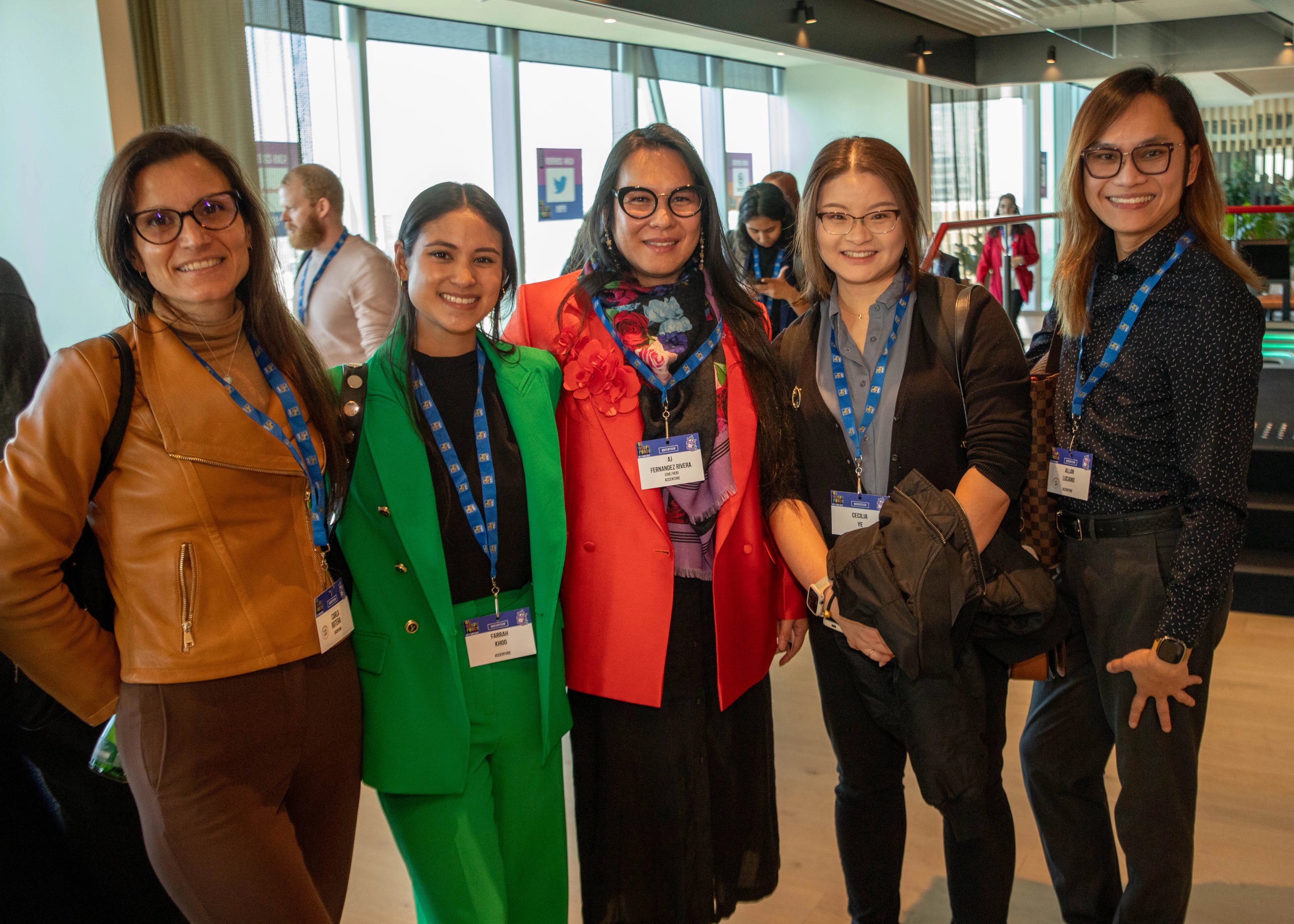 A group of five smiling people in business attire stands together indoors at a networking event. They wear name badges and blue lanyards, exuding a cheerful and friendly atmosphere.