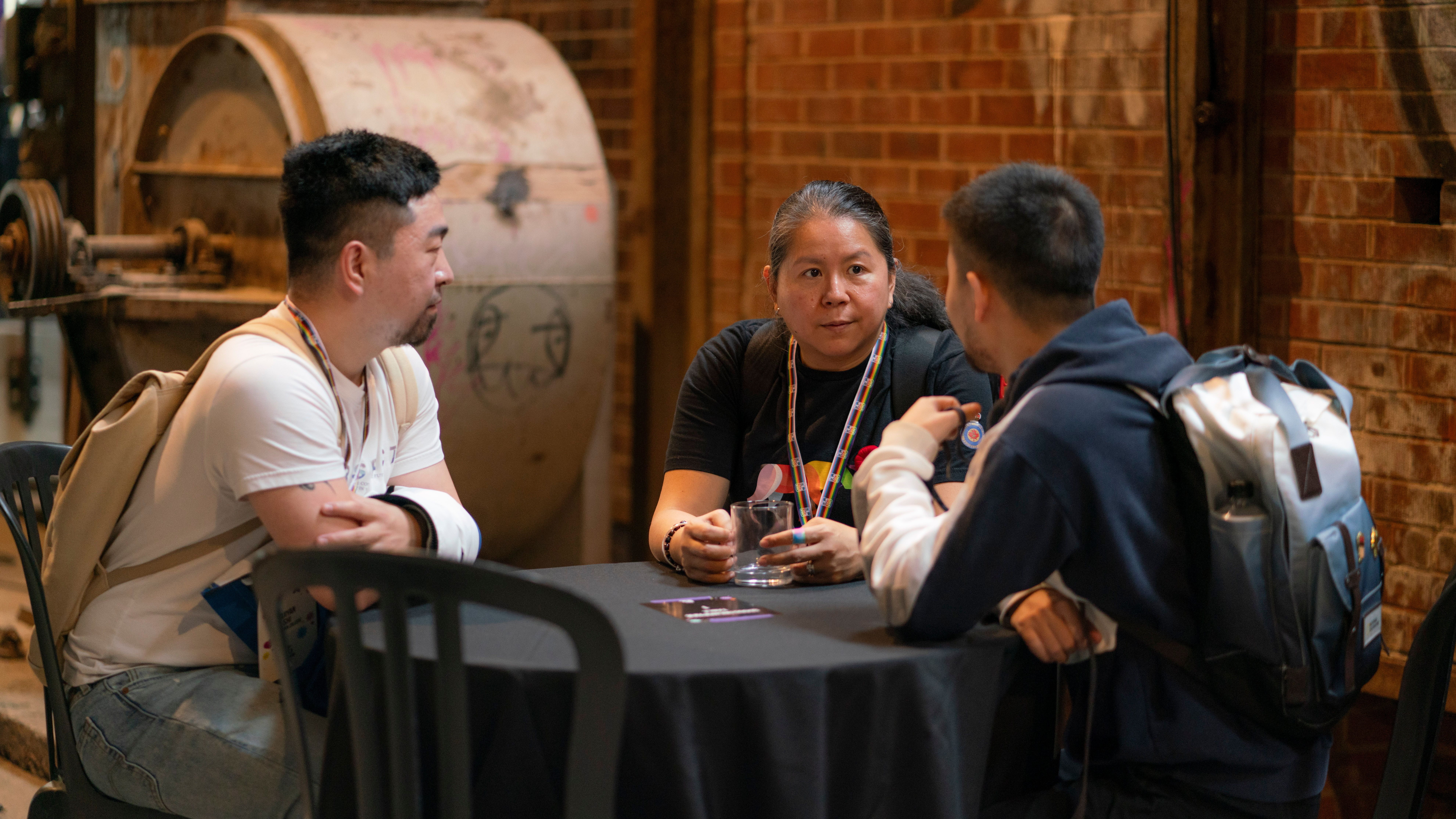 Three people sit around a black table engaged in conversation. They appear focused. A brick wall and industrial equipment are in the background.