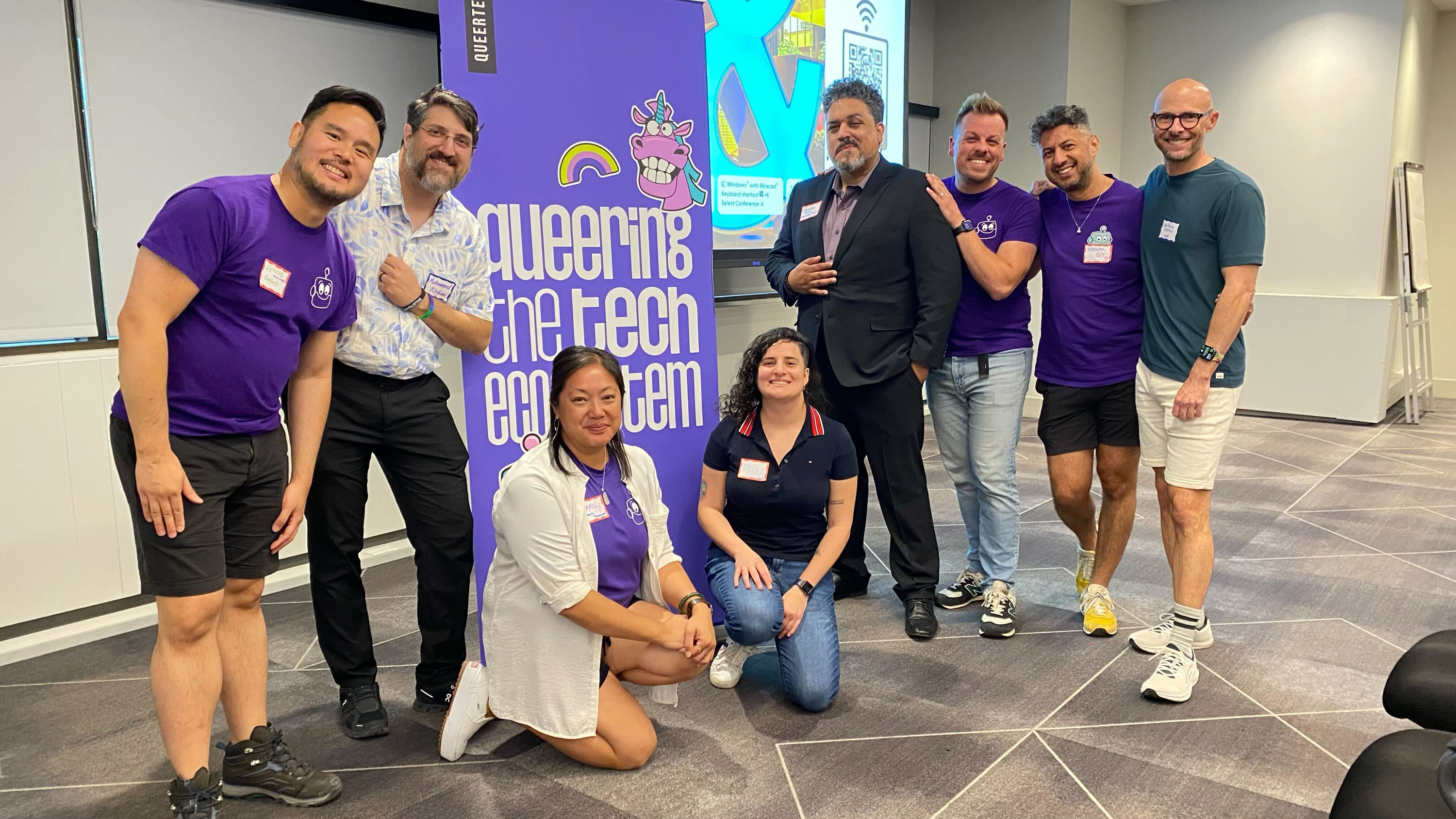 A diverse group of eight people poses cheerfully in front of a banner that reads "Queering the Tech Ecosystem," conveying a tone of inclusivity and community.