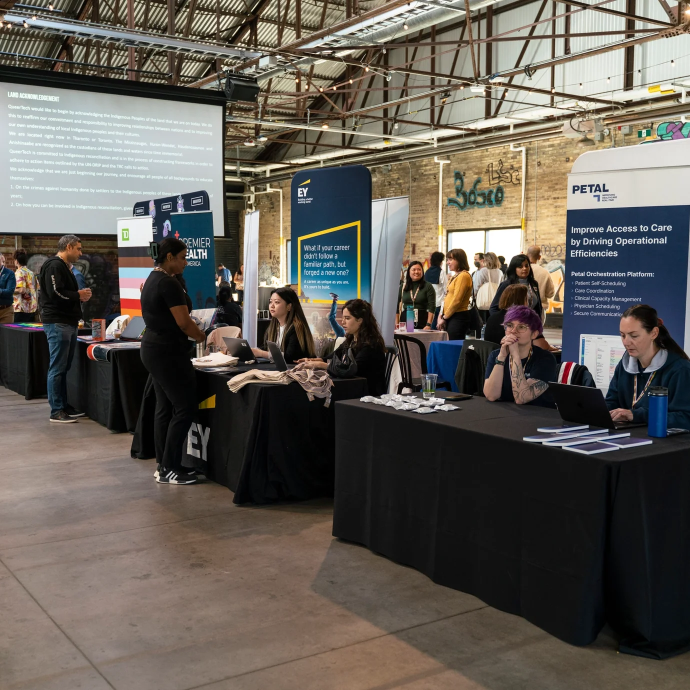 Indoor career fair with multiple booths featuring company banners. People interact and exchange information. The atmosphere is lively and professional.