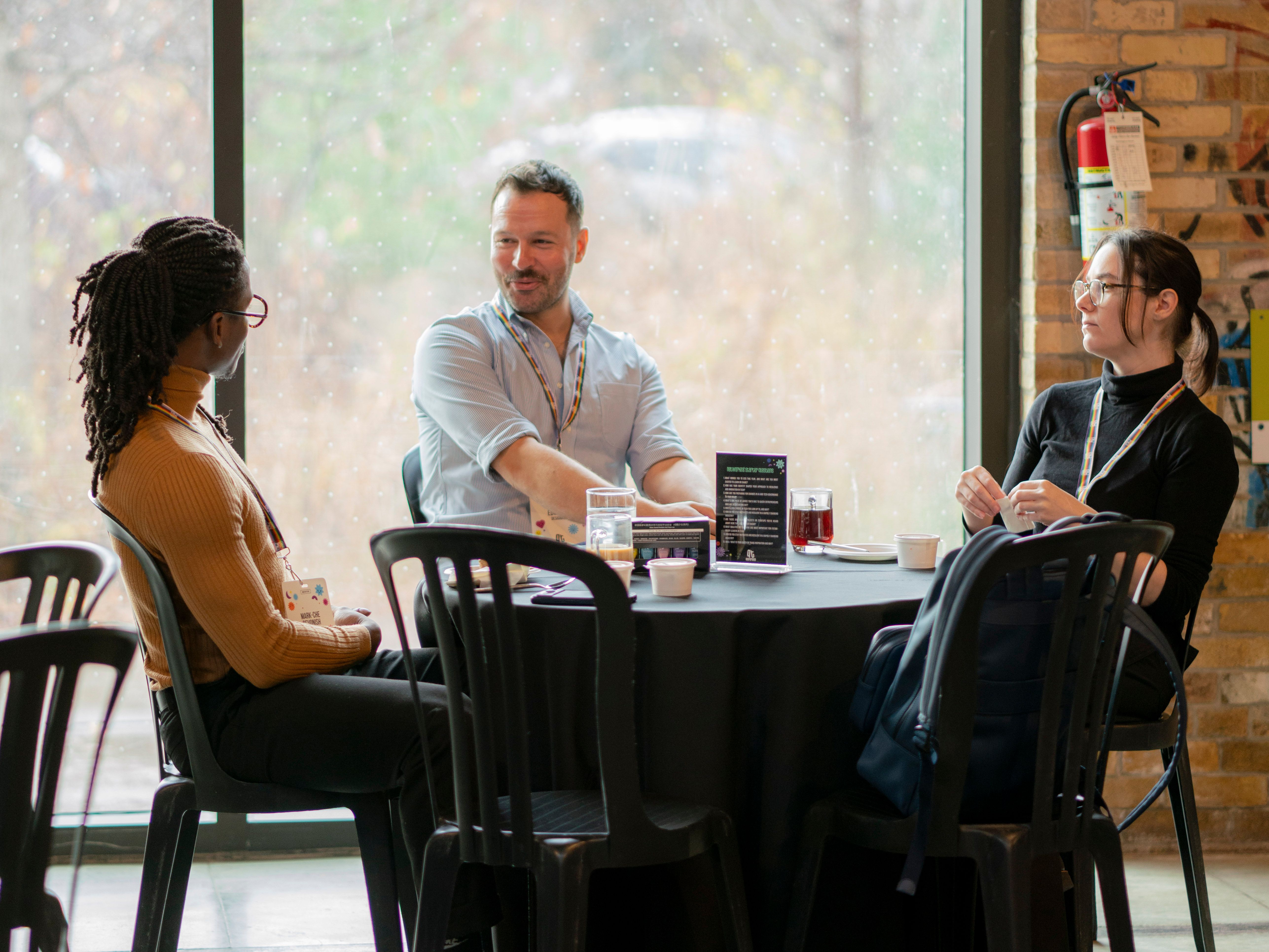 Three people sit around a café table in discussion, with a large window showing a blurred outdoor scene. The mood is relaxed and engaged.