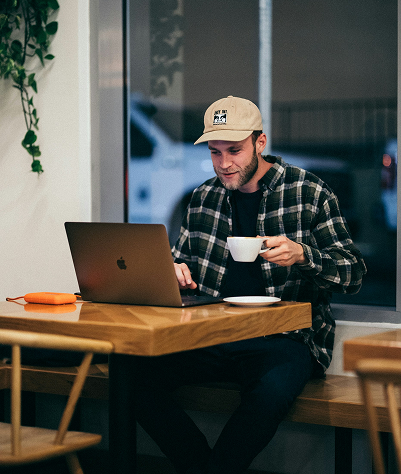 A man in a plaid shirt and cap smiles while using a laptop and holding a coffee cup in a cozy café. A plant hangs nearby, adding warmth to the scene.