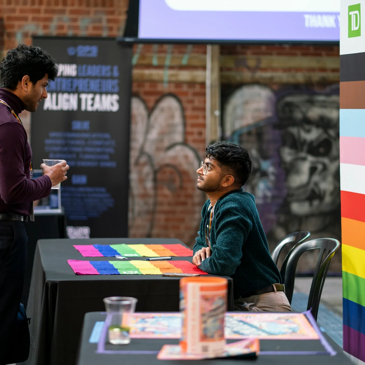 Two men converse at a table covered with rainbow paper. One stands while the other sits, creating an atmosphere of engagement. Graffiti decorates the brick wall.