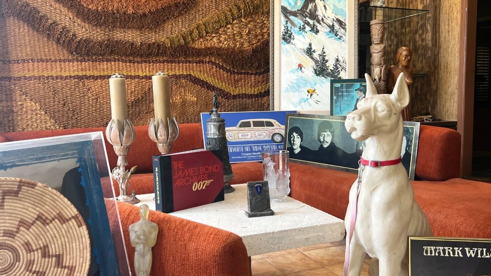 Toluca Lake Hollywood insider estate sale room display with orange sofa, woven tapestry, Beatles photograph, James Bond book, art, and white dog statue.