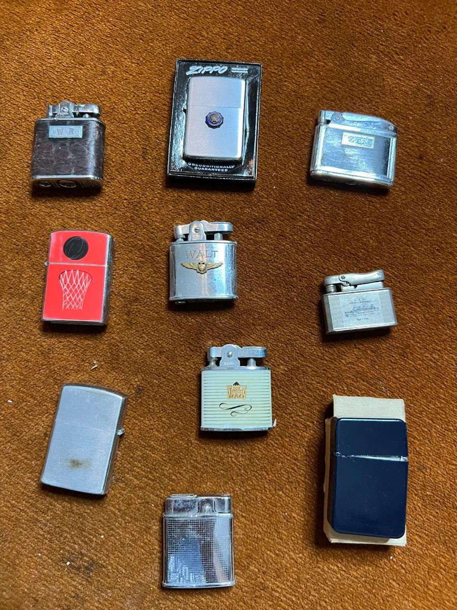 Collection of vintage lighters including Zippo-style and pocket lighters displayed on brown fabric.