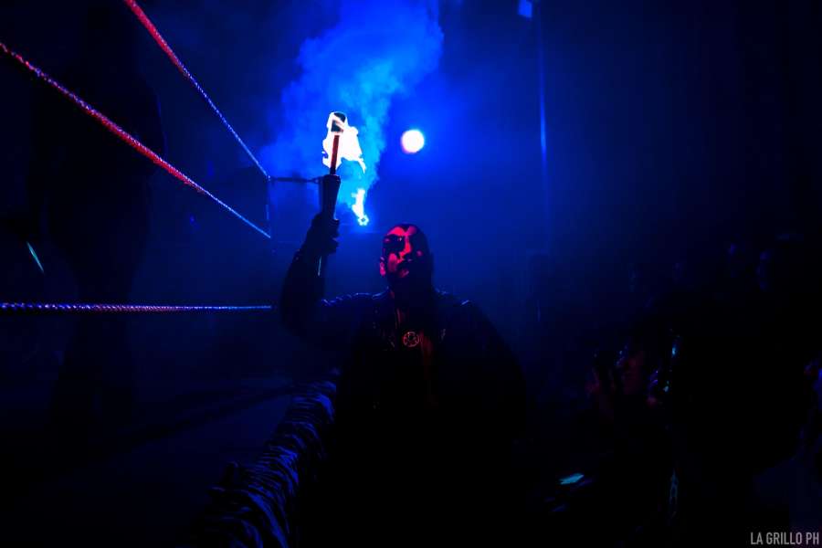 Azagoth caminando entre el ring y los asistentes a poca luz con una antorcha en la mano