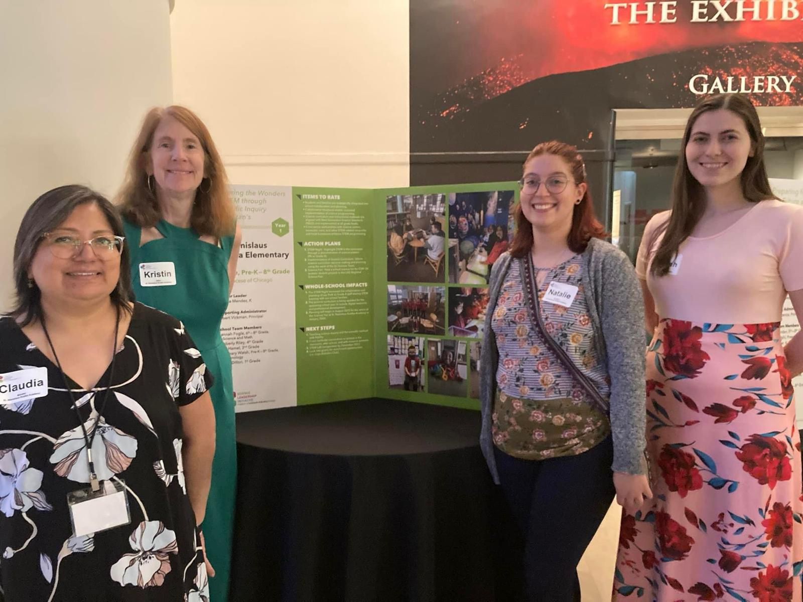 4 teachers stand around a table that has a tri-fold poster board. 