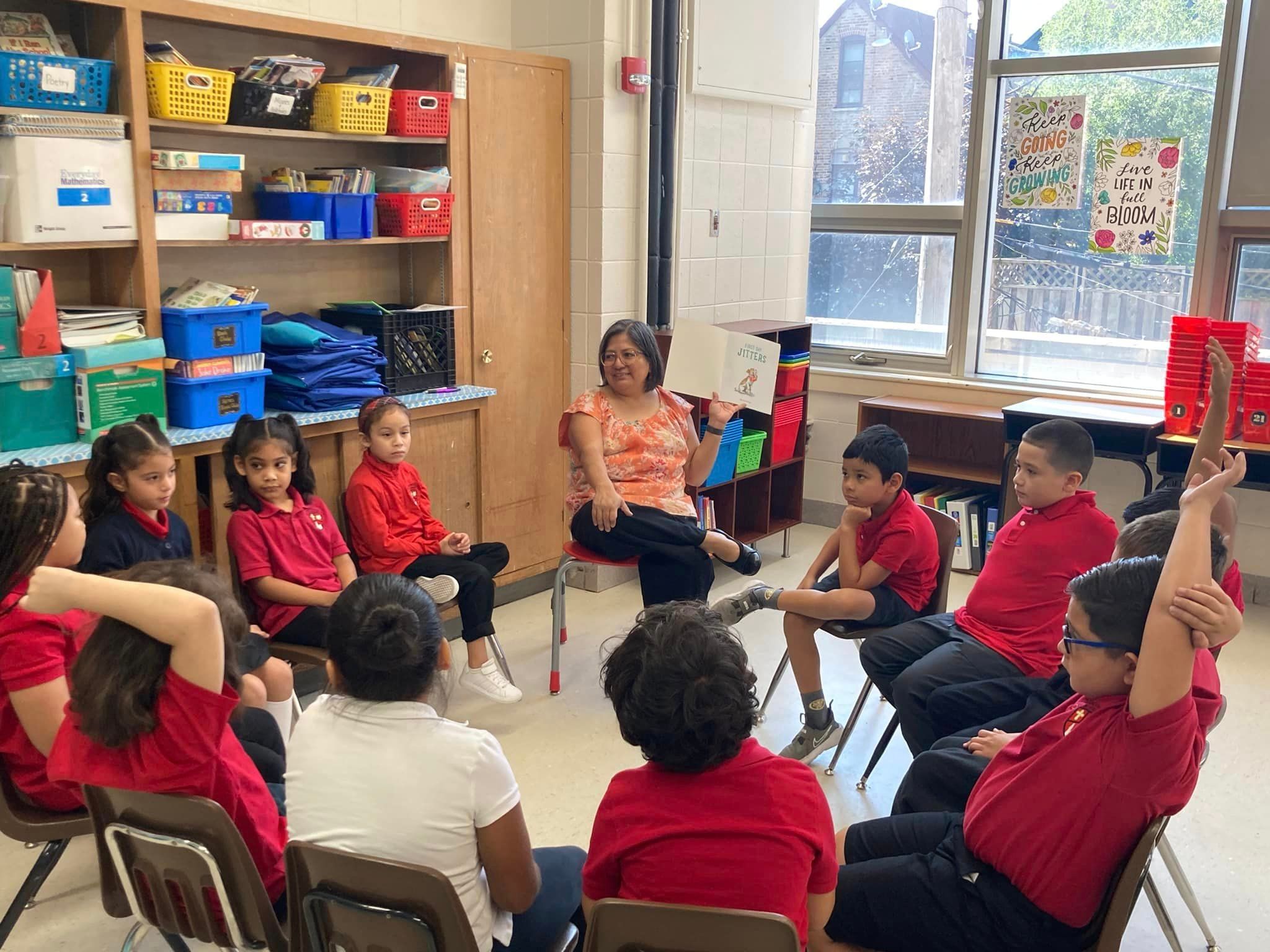 Almost a dozen students sit in chairs positioned all to form a circle. Claudia sits in a chair with a book open in front of her.