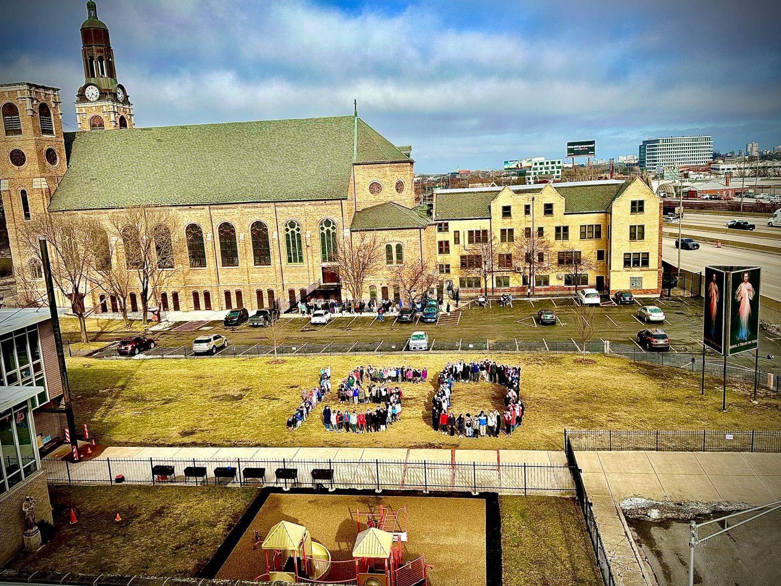 aerial view of a grassy field in front of a church. Dozens of people stand in certain spots in order to form the number "150".