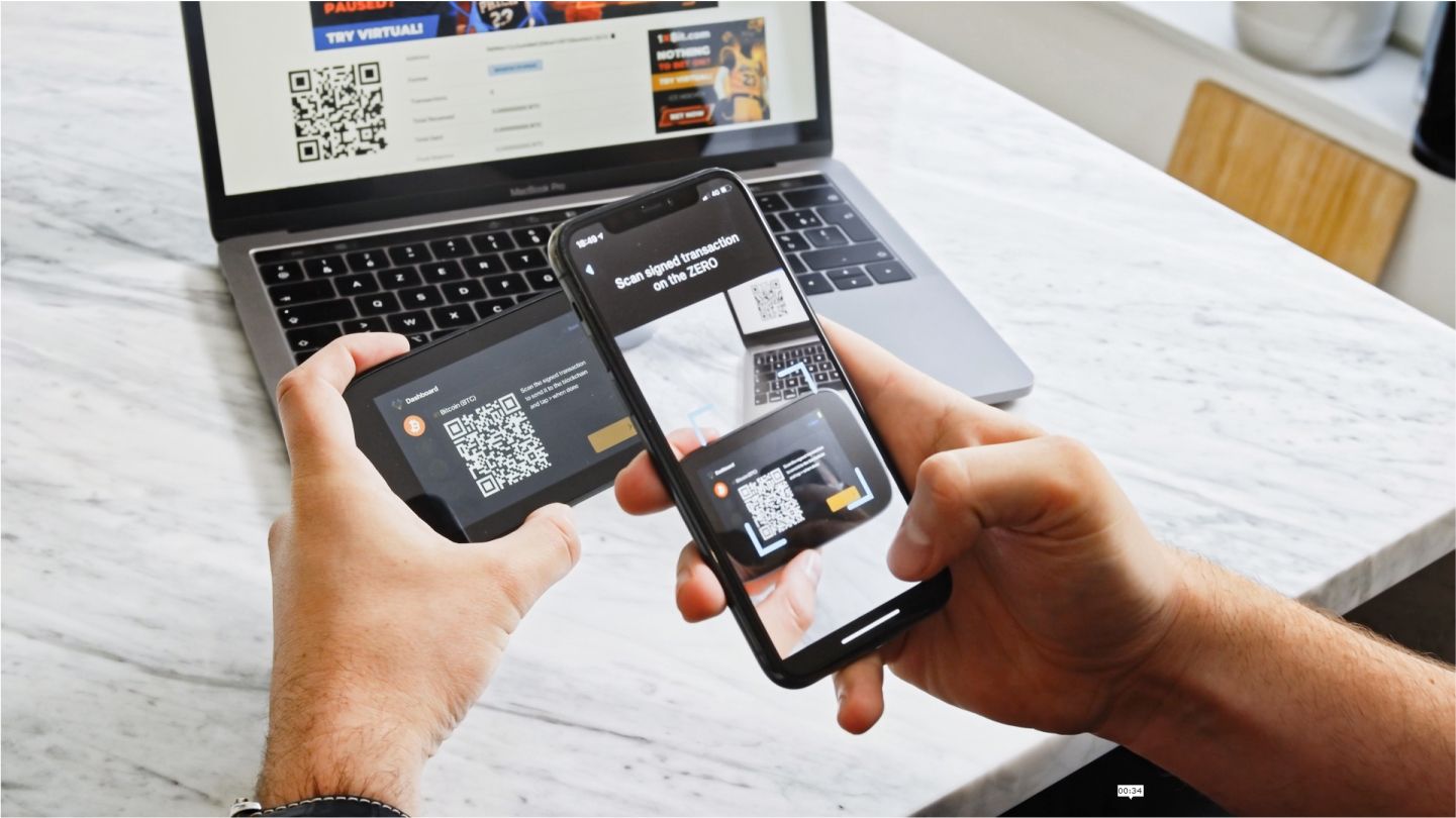 Two hands holding a smartphone that scans a QR code on a small device screen, with an open laptop softly blurred in the background.