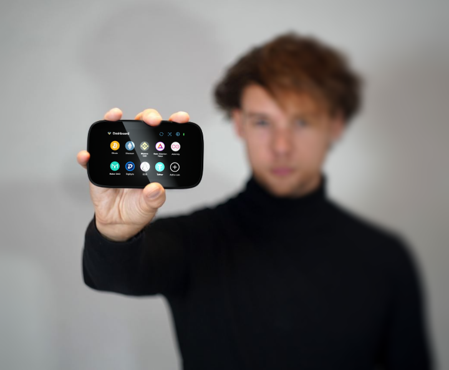 A person in a dark sweater holding a small hardware wallet whose screen displays a neat list of coin icons.