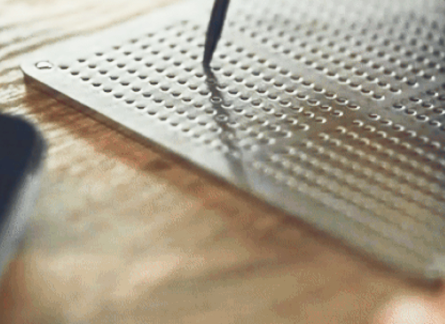 Close-up of a stainless steel backup plate marked with rows of punched holes, with a metal stylus resting beside it.