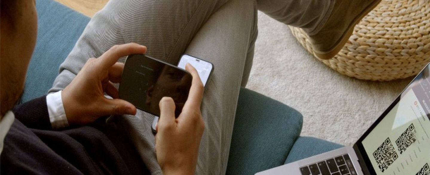 A person relaxing on a sofa holding a small hardware wallet in one hand, with an open laptop resting on the cushion beside them.