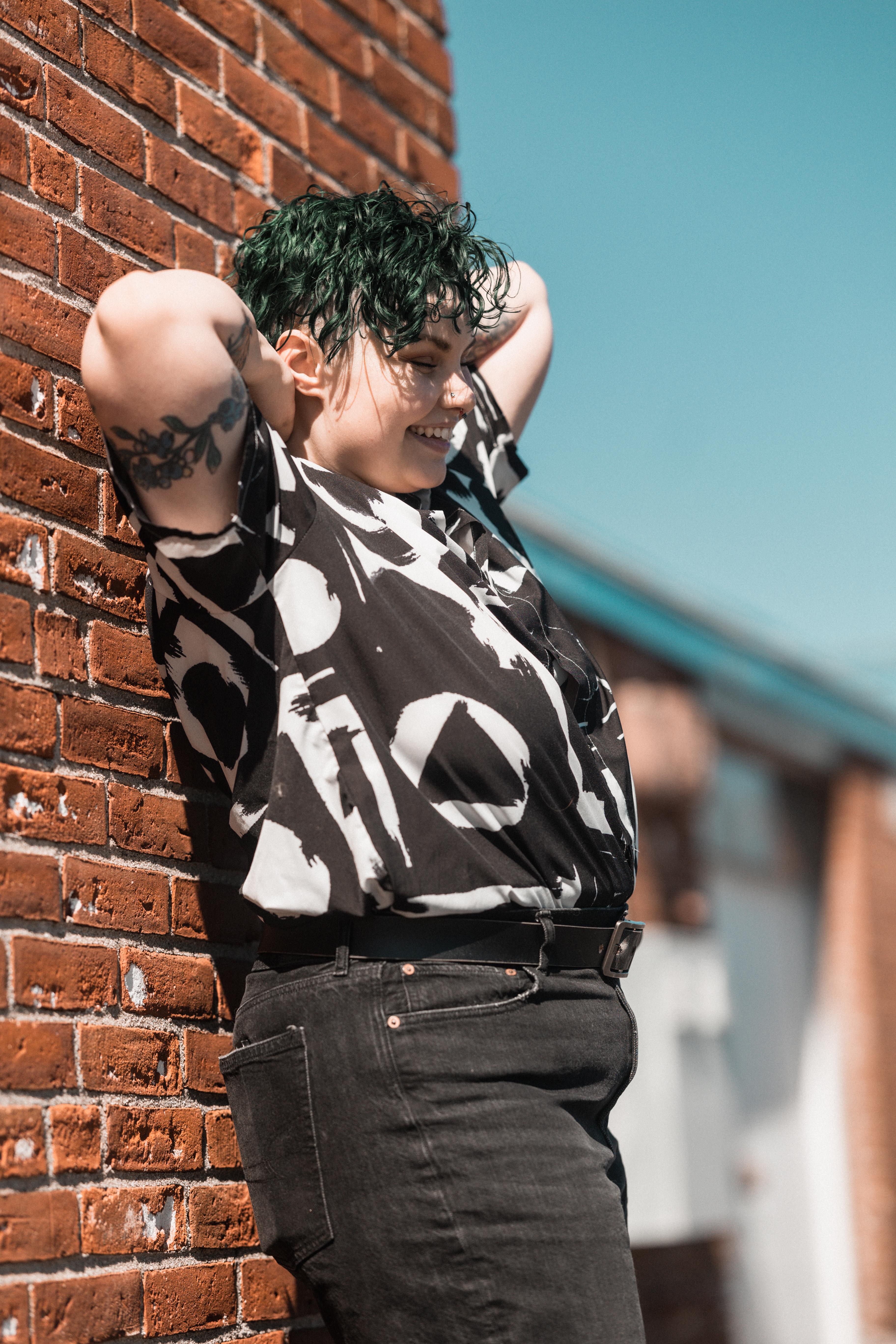 Kyah with their back against a brick wall and their arms behind their head, laughing. Photo credit WCKD collective.