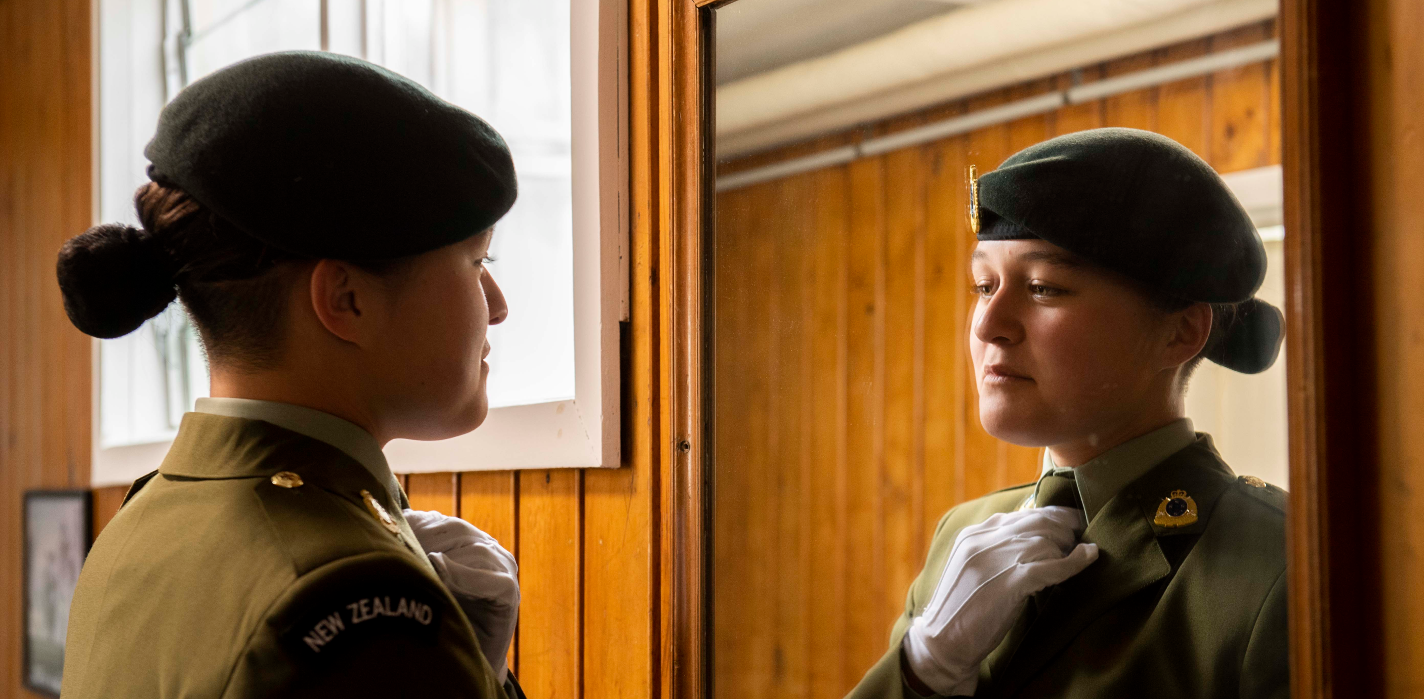 A female New Zealand Defence Force soldier in dress uniform adjusts her tie while looking at her reflection in a mirror. The image captures her from the side and in the mirror, showing her focused expression and attention to detail in preparing her uniform. 