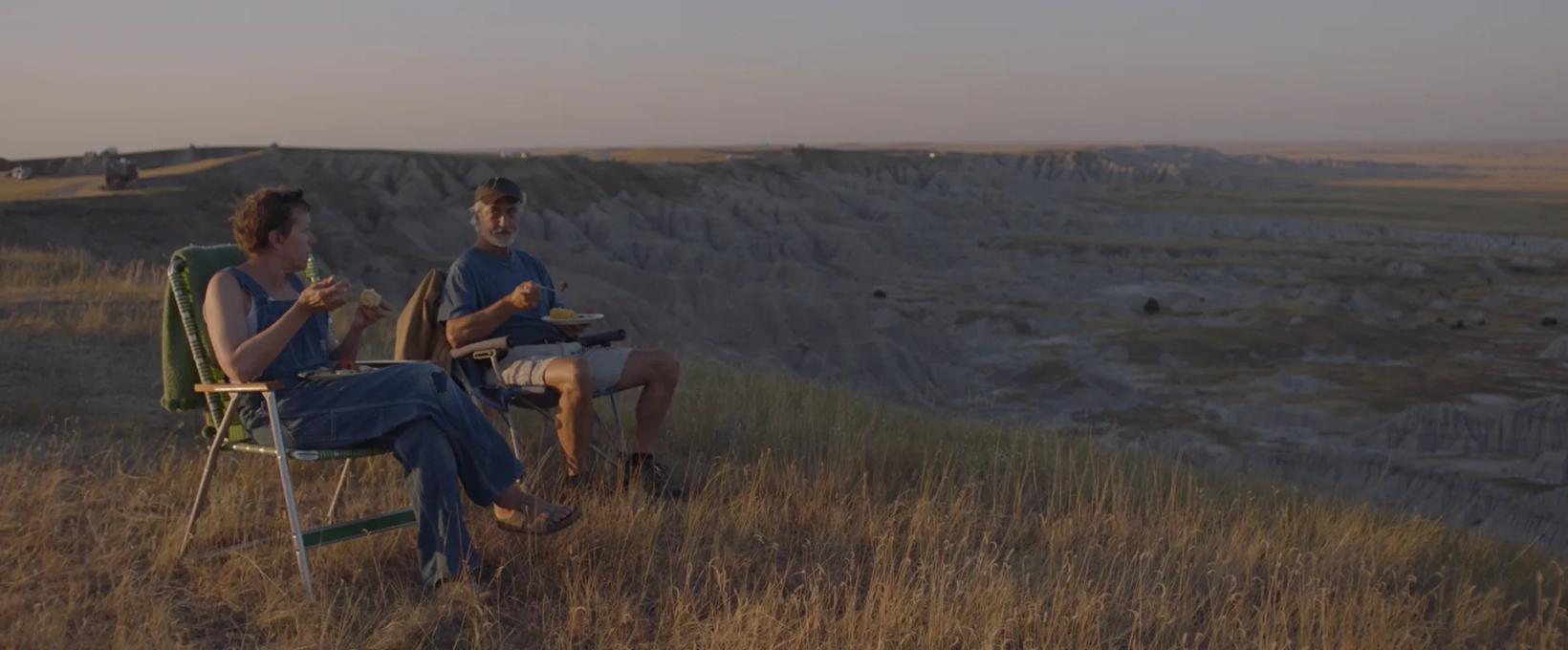 a couple of men sitting in chairs on a grassy hill overlooking a valley