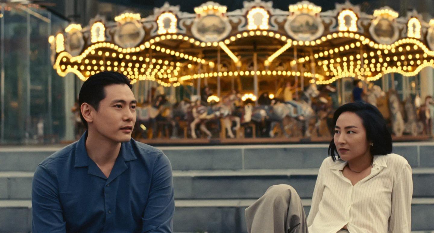 Teo Yoo, Greta Lee sitting in front of a carousel