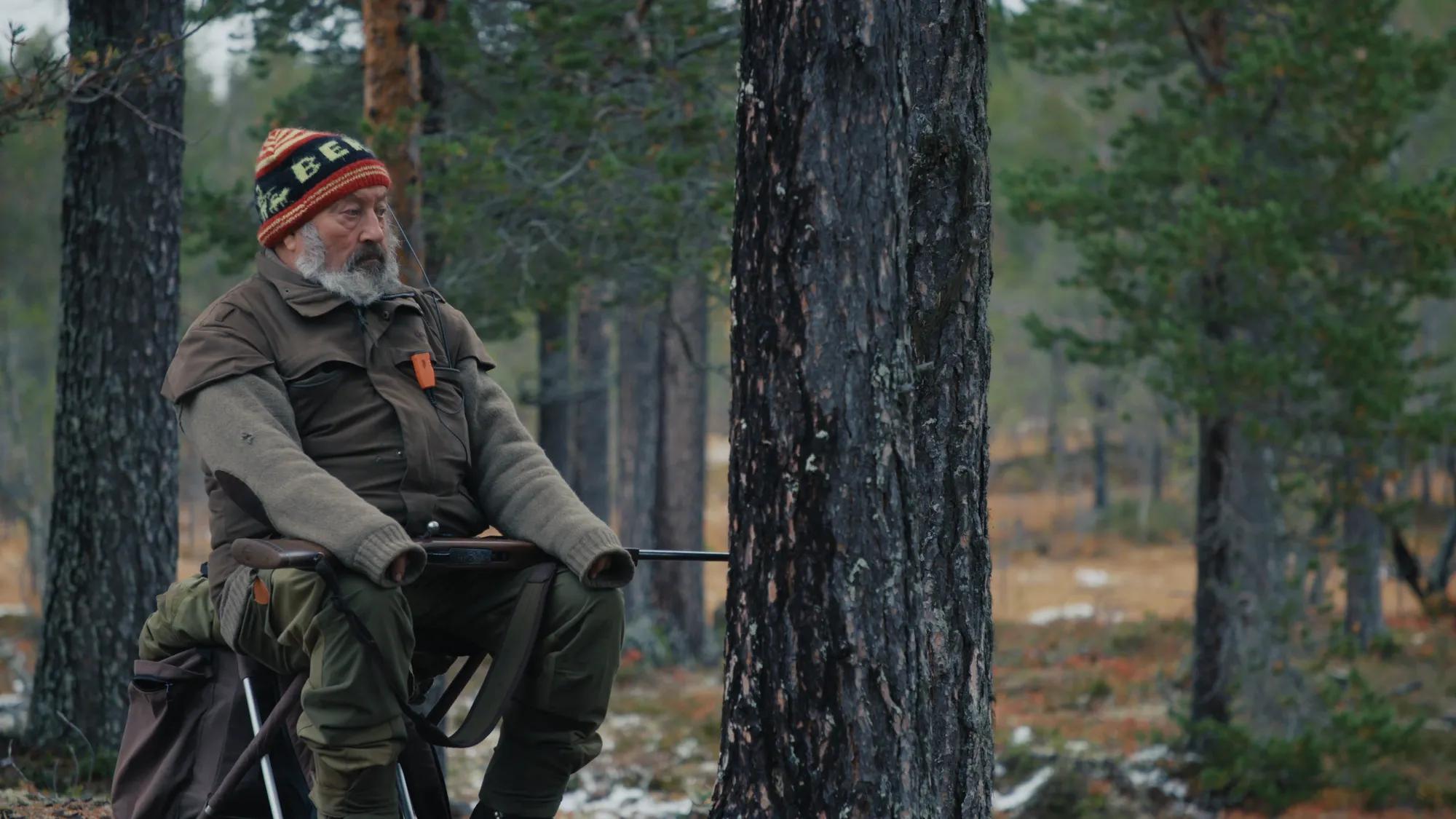a man with a beard and hat sitting on a chair in the woods
