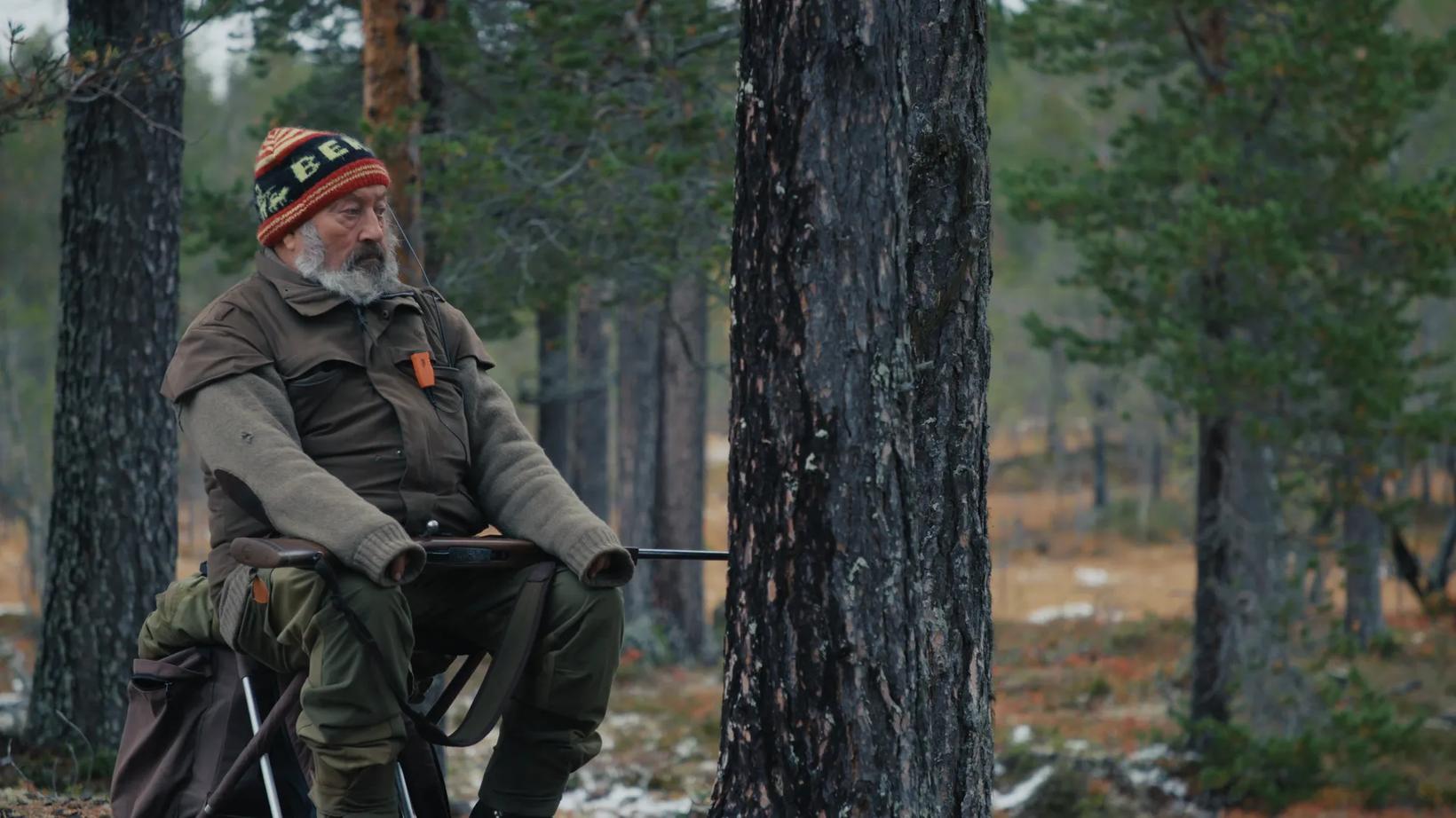 a man with a beard and hat sitting on a chair in the woods