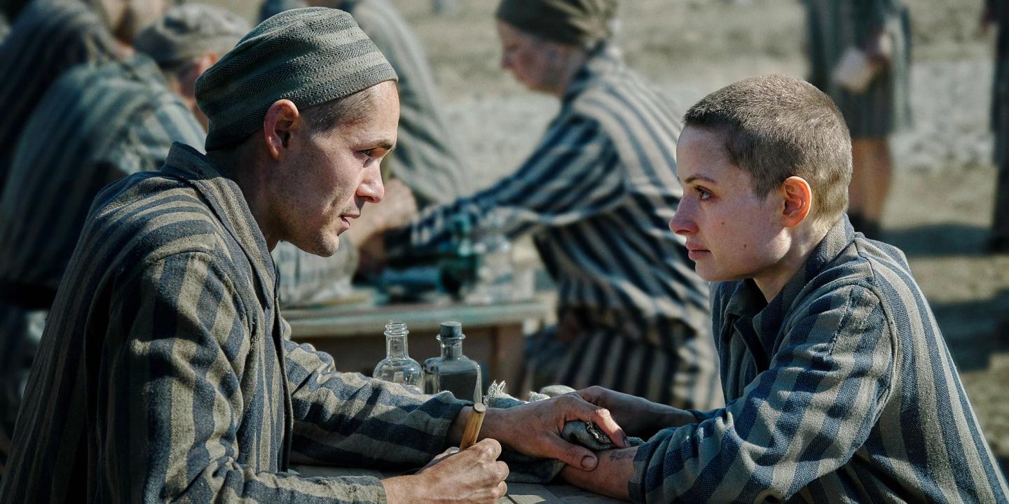 Jonah Hauer-King og Anna Próchniak ser på hverandre mens han tatoverer henne, begge iført fangeuniformer, i serien Tatovøren fra Auschwitz