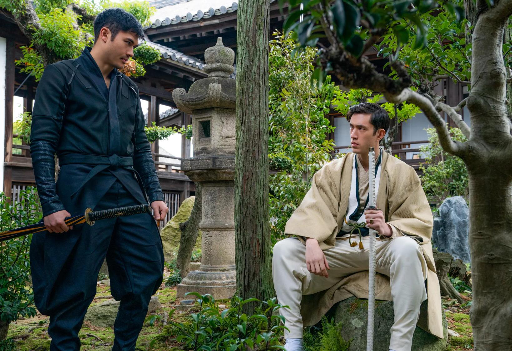 Andrew Koji sitting on a stool next to a person holding a rifle