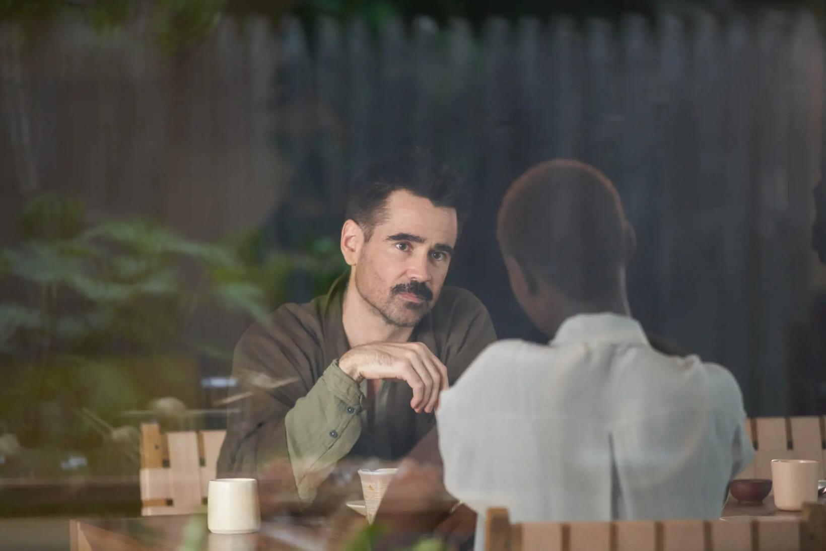 Colin Farrell sitting at a table with a woman behind him