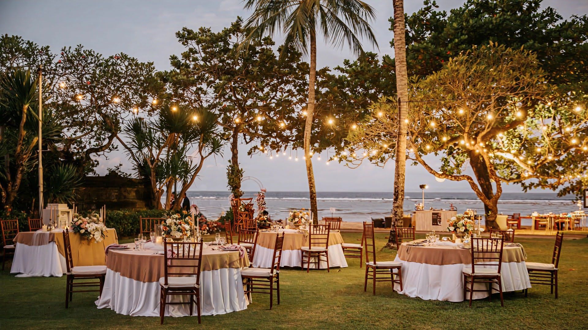 Beachfront wedding reception with round tables, string lights, and ocean view at dusk.