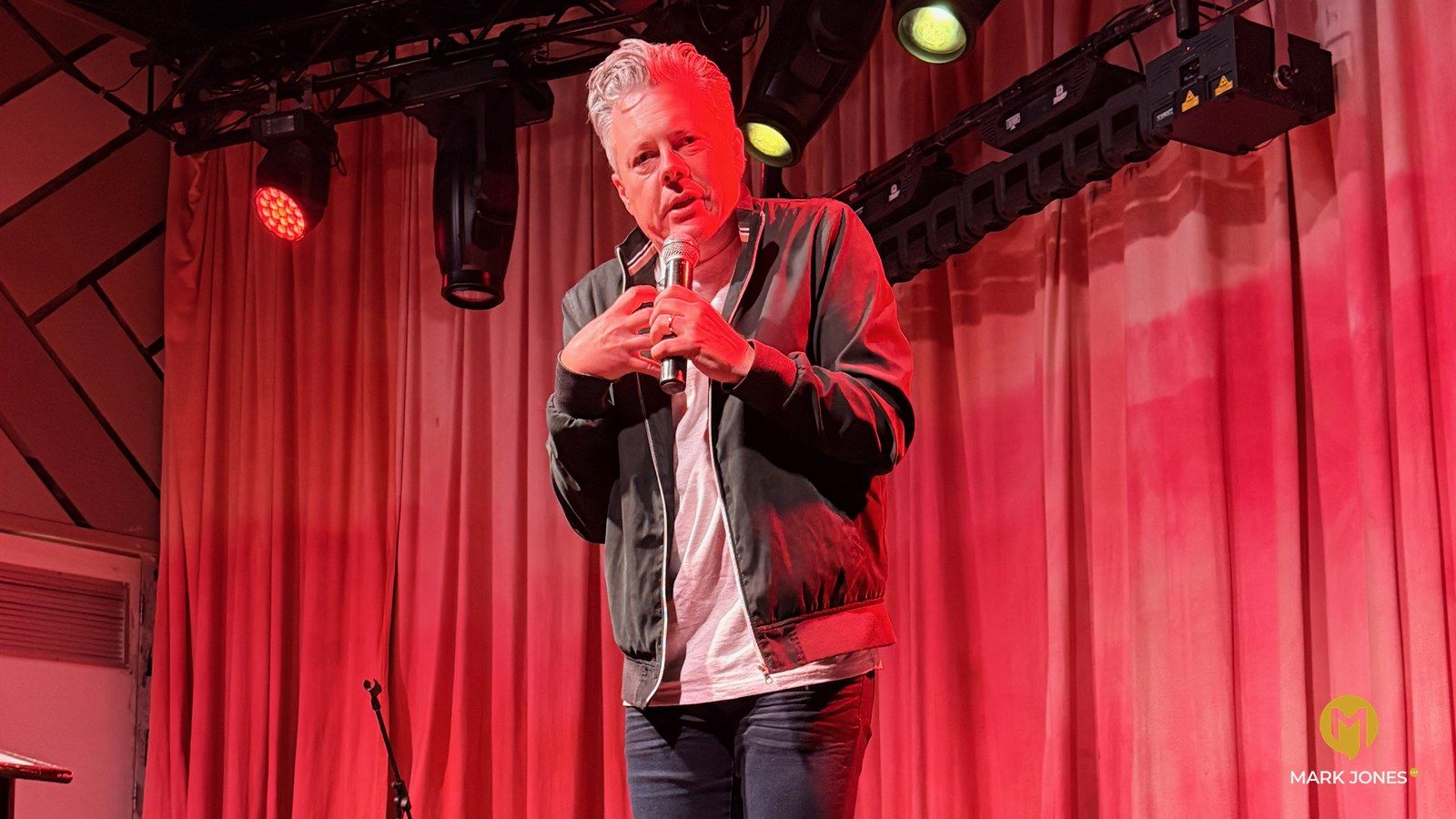 Mark Jones on stage at His Story in Sydney, holding a microphone to his chest under red stage lights