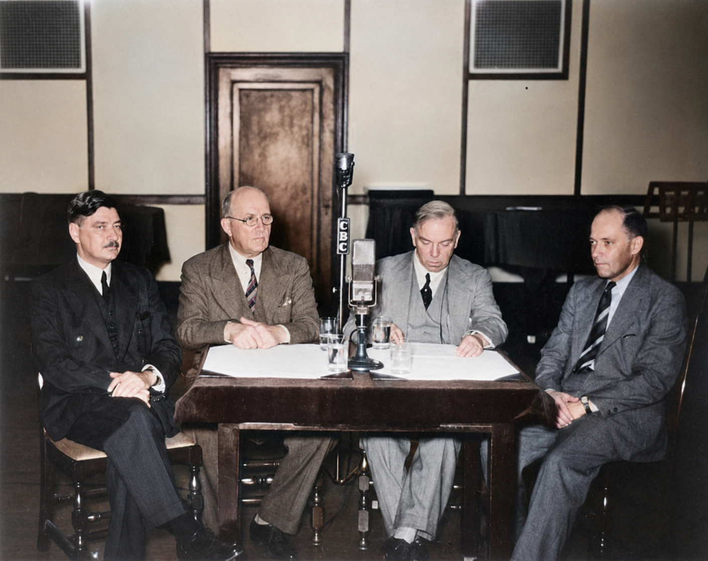 Mackenzie King and three Cabinet ministers sit around a table with papers and two radio microphones during an Ottawa broadcast.