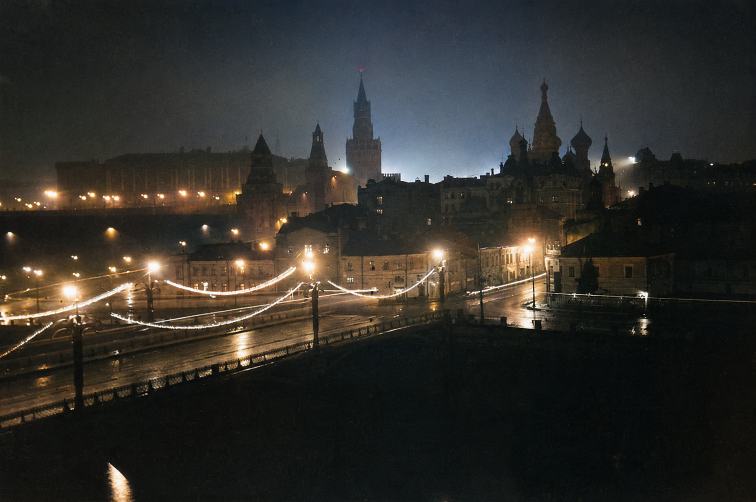 The floodlit walls and towers of the Kremlin and the clustered domes of St Basil's Cathedral rise above the Moskva river at night, their lights mirrored in the dark water below.