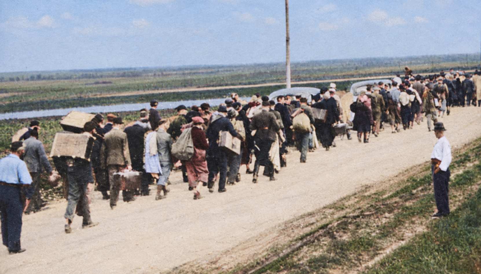 A long column of civilians walking away from the camera along a dirt road across open farmland, carrying suitcases, bundles and sacks, with a covered cart among them and a man standing at the roadside watching them pass.