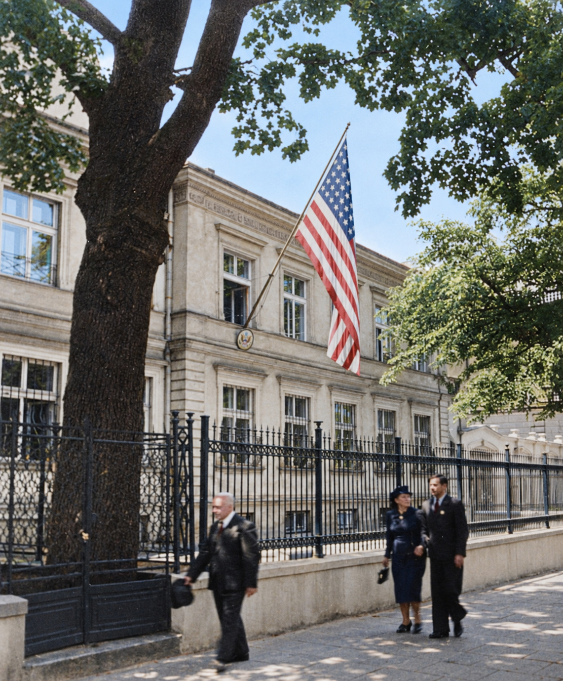 Street view of the US Embassy chancery in Warsaw behind railings, with an American flag above the pavement and civilians nearby.