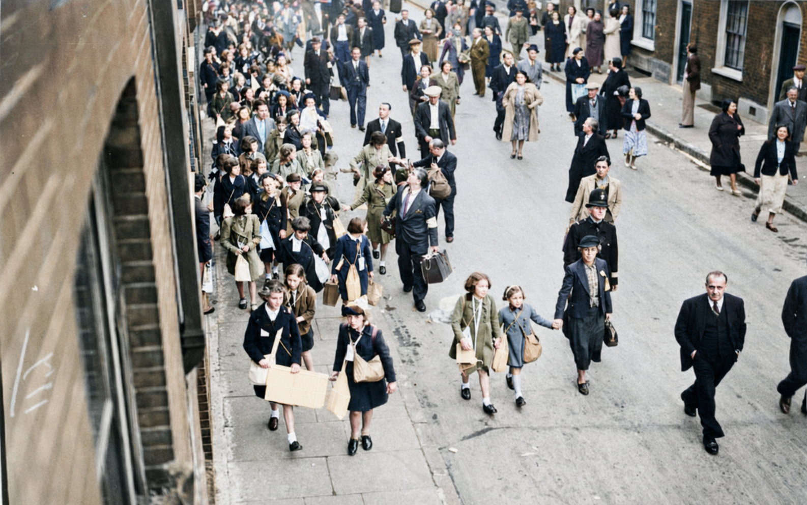 Children carrying suitcases and small cardboard boxes on strings, walking in a line beside adult marshals.