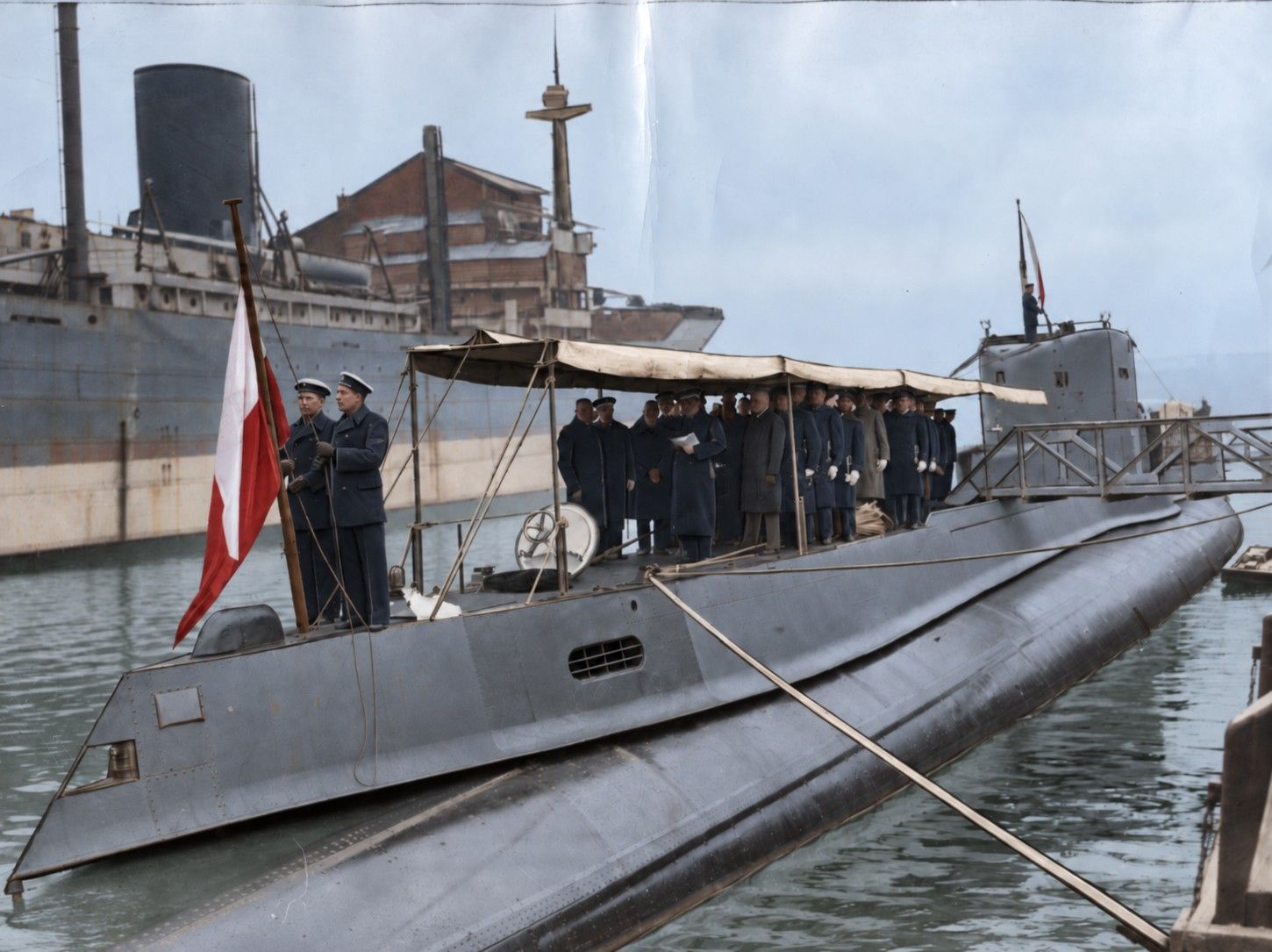 The submarine Orzel lies alongside at the Vlissingen shipyard during a ceremonial raising of the ensign, with sailors and officials on deck.