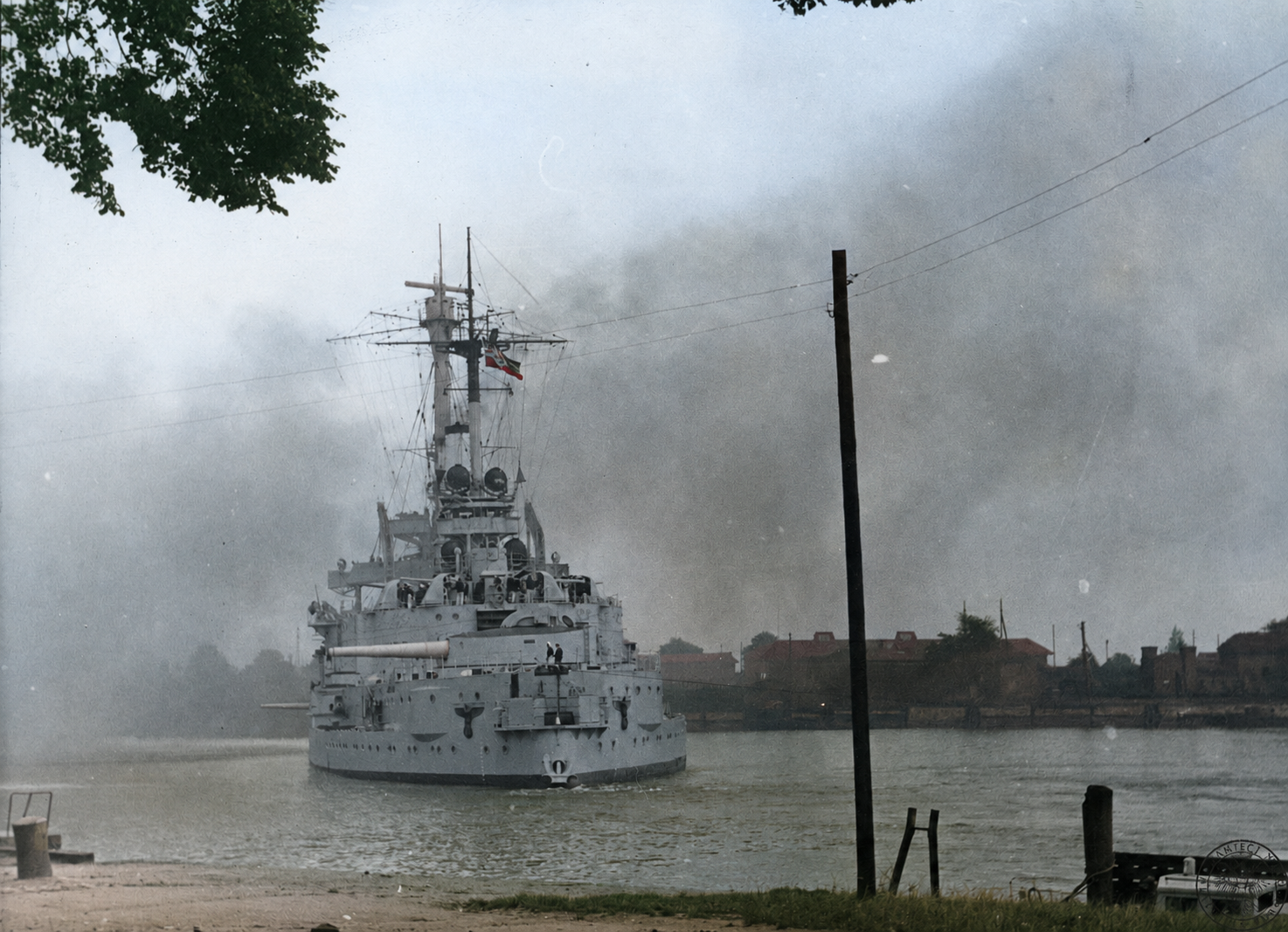 A battleship lying close to a low riverbank, with smoke rising beyond the far shore.