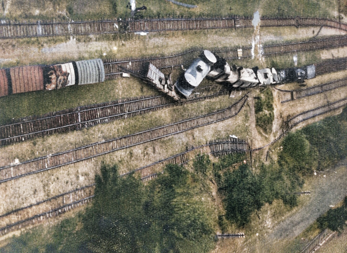 Aerial view of a railway junction with several sets of tracks, where a derailed armoured train lies buckled across the rails, its locomotive and wagons thrown at angles beside a siding.