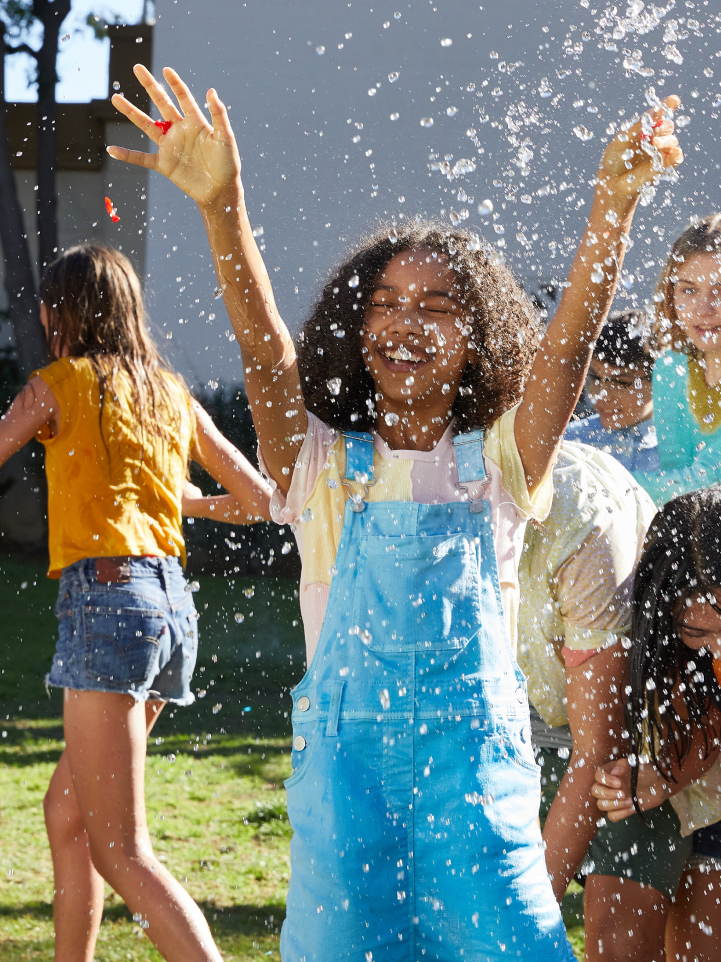 Joyful children playing with water on a sunny day, with a smiling girl in denim overalls raising her hands up in the air, enjoying the water splashes, while other kids are seen in the background, capturing a moment of pure summer fun.
