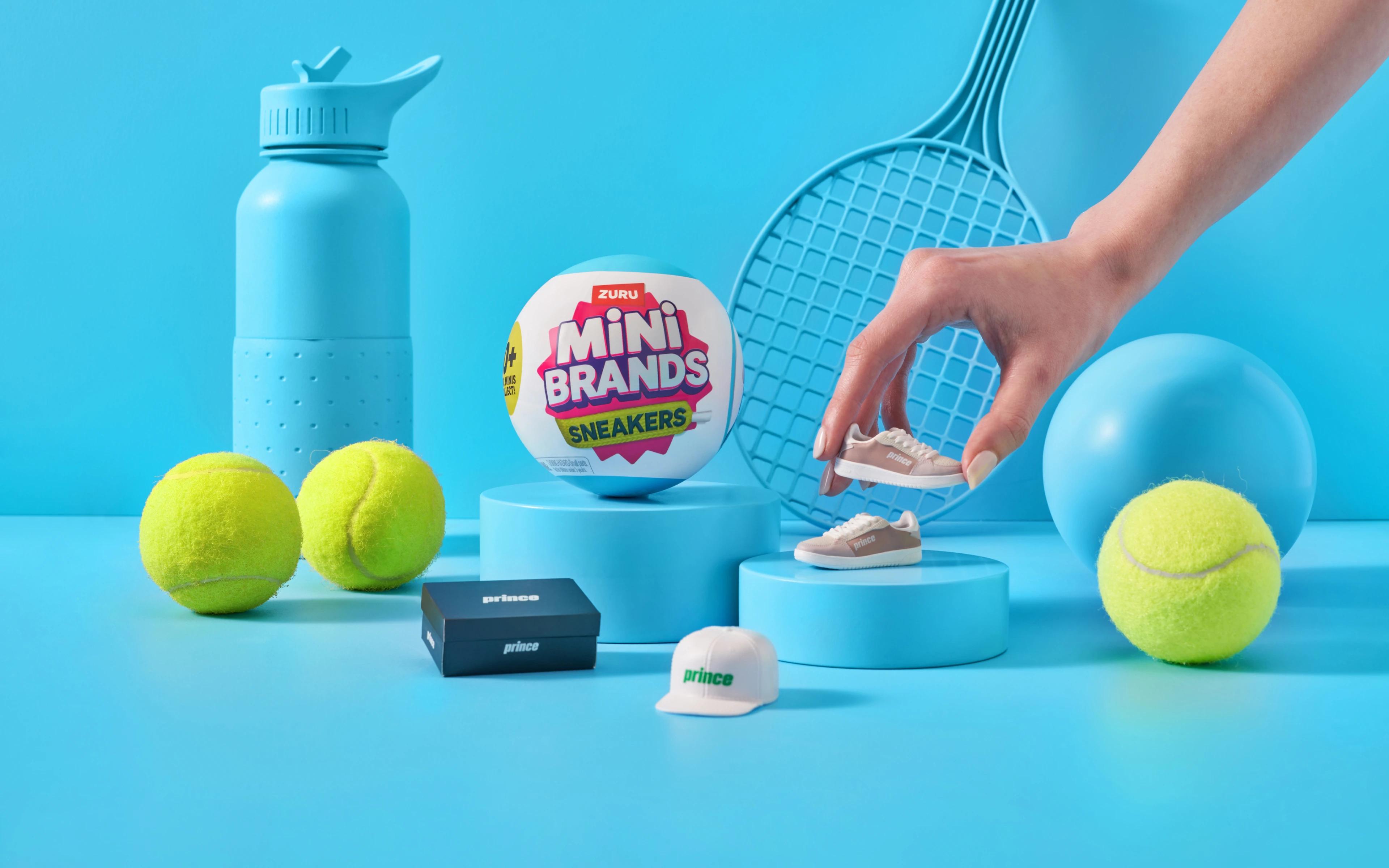 Interactive setup of ZURU Mini Brands Sneakers collection with a hand placing a miniature sneaker on a pedestal, surrounded by sports-themed items including tennis balls, a water bottle, a cap, and a racket on a blue background.