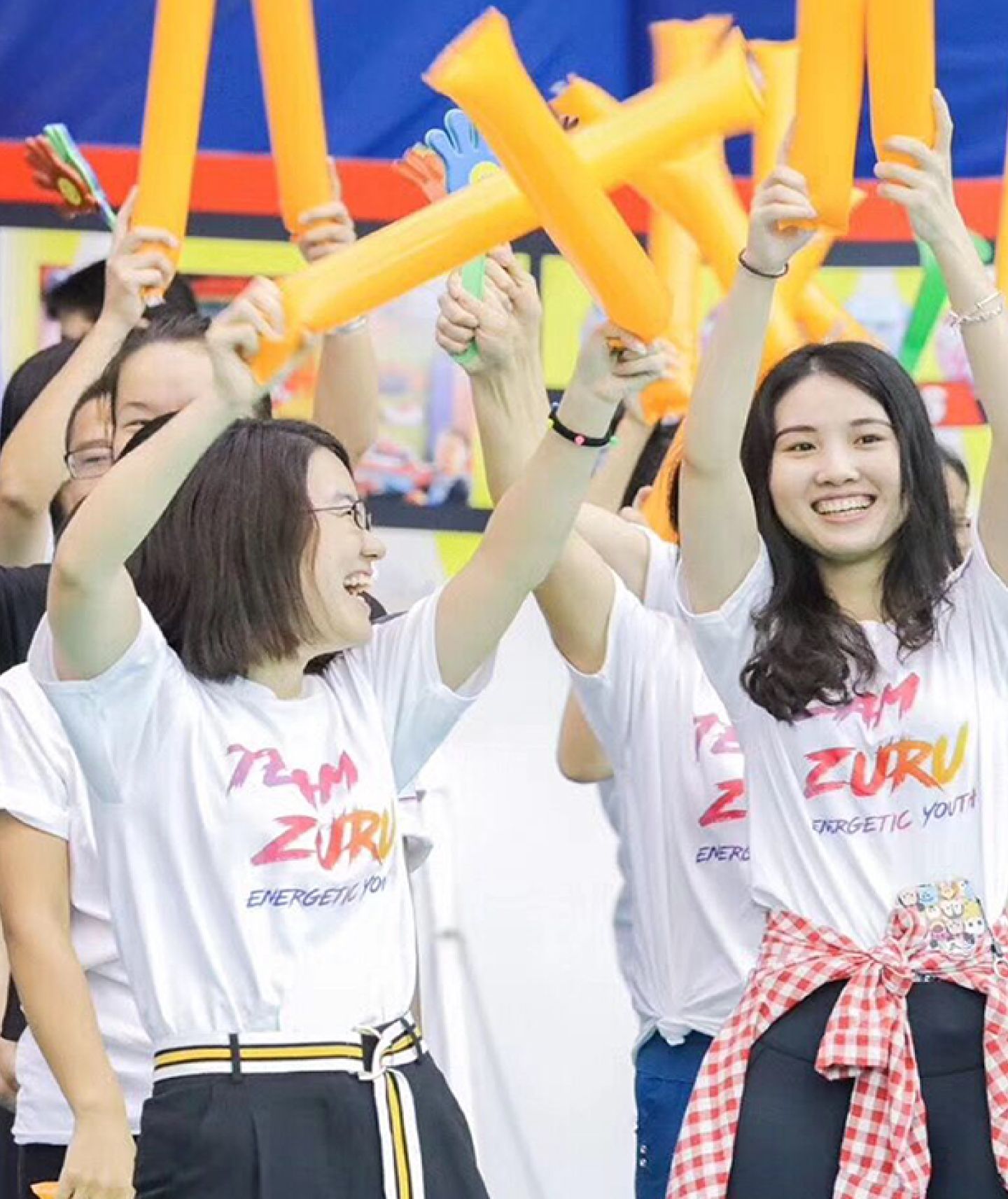 Exuberant young adults at a team event, wearing 'Team ZURU' t-shirts and cheerfully waving orange inflatable sticks, with expressions of excitement and camaraderie, set against a backdrop of a festive crowd.