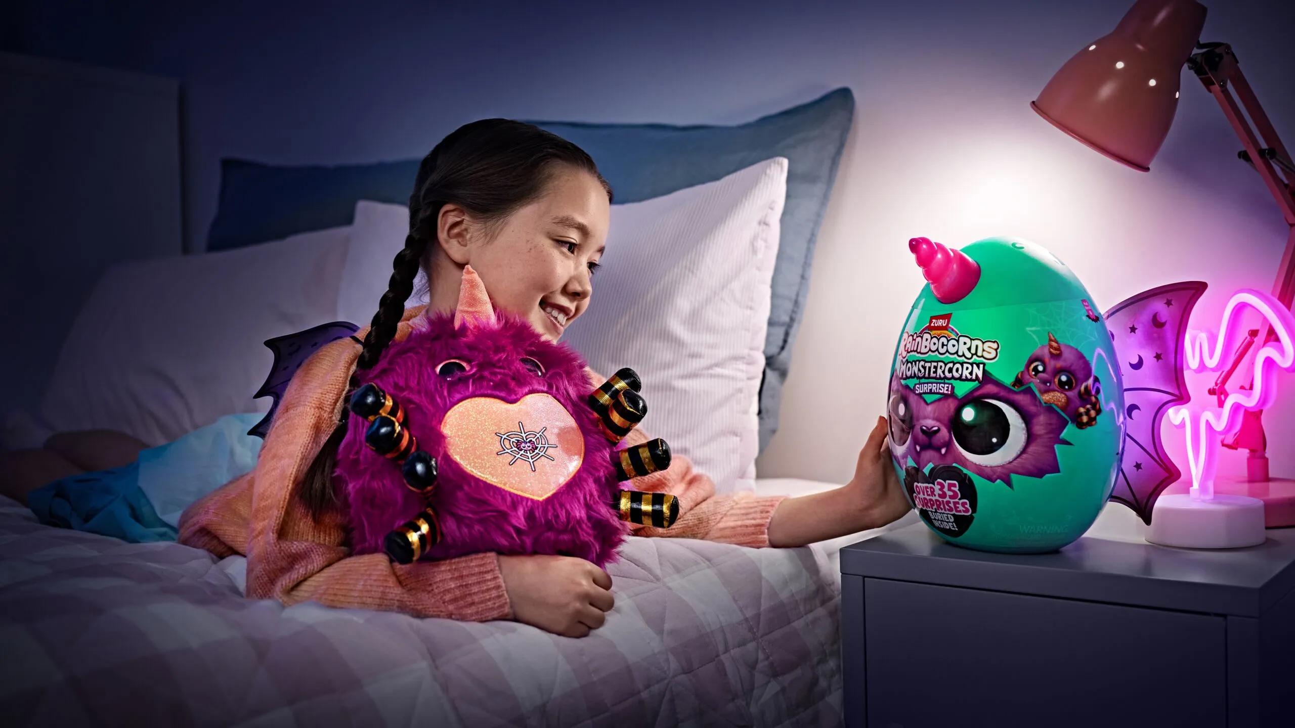 A young girl smiles while holding a fluffy purple Rainbocorns Monstercorn plush toy and reaching towards a colorful Monstercorn Surprise egg.
