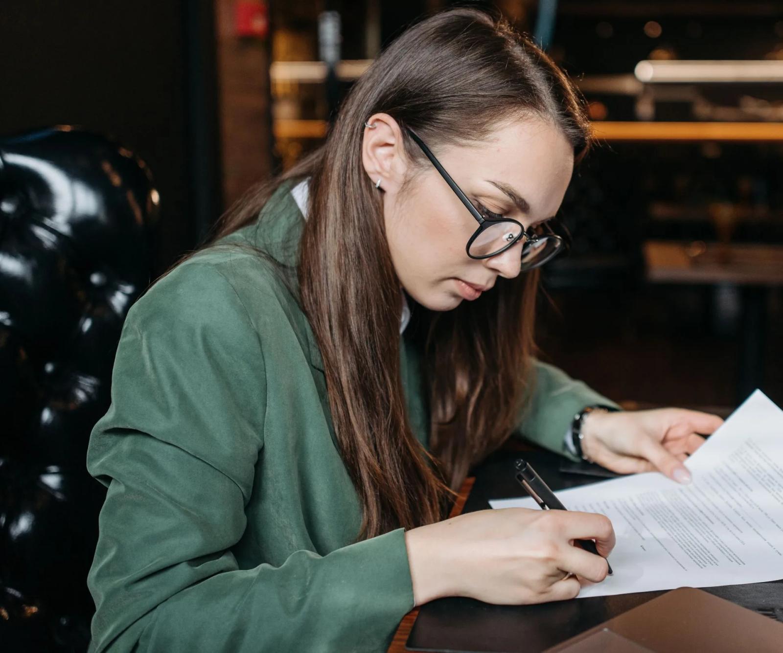 Una mujer con gafas y chaqueta verde firma un documento en su escritorio.