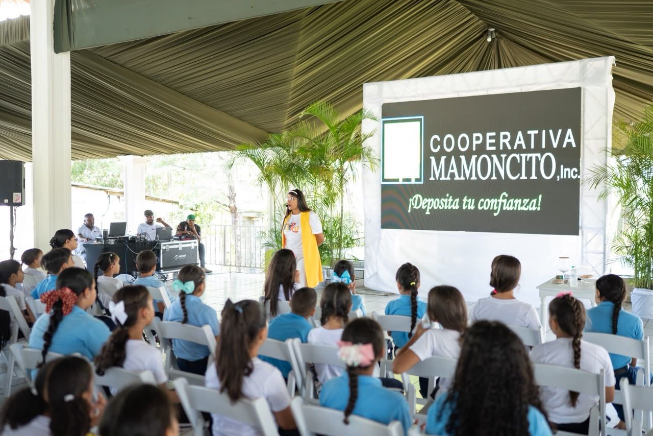 Niñas y niños con uniforme escolar bajo la carpa de Mi primera alcancía, frente al cartel de Cooperativa Mamoncito.