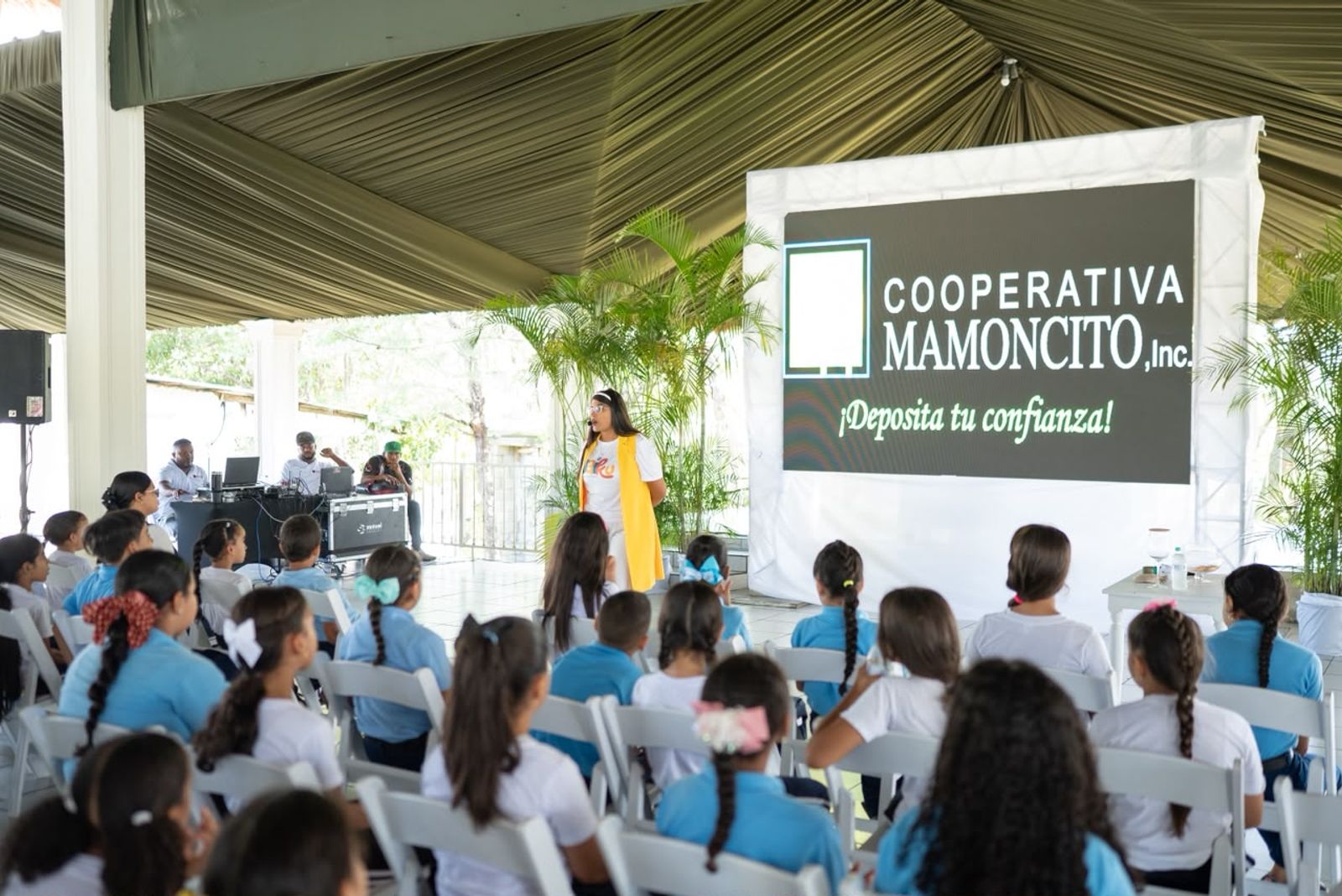 Niñas y niños con uniforme escolar bajo la carpa de Mi primera alcancía, frente al cartel de Cooperativa Mamoncito.