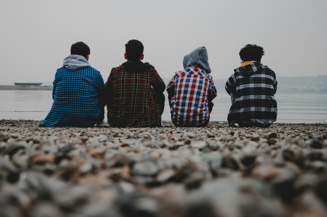 Four young men sitting on a rocky shore, facing the water.