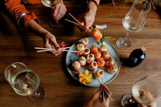 Four people reach in with chopsticks to share a plate of salmon sushi rolls on a wooden table set with wine glasses