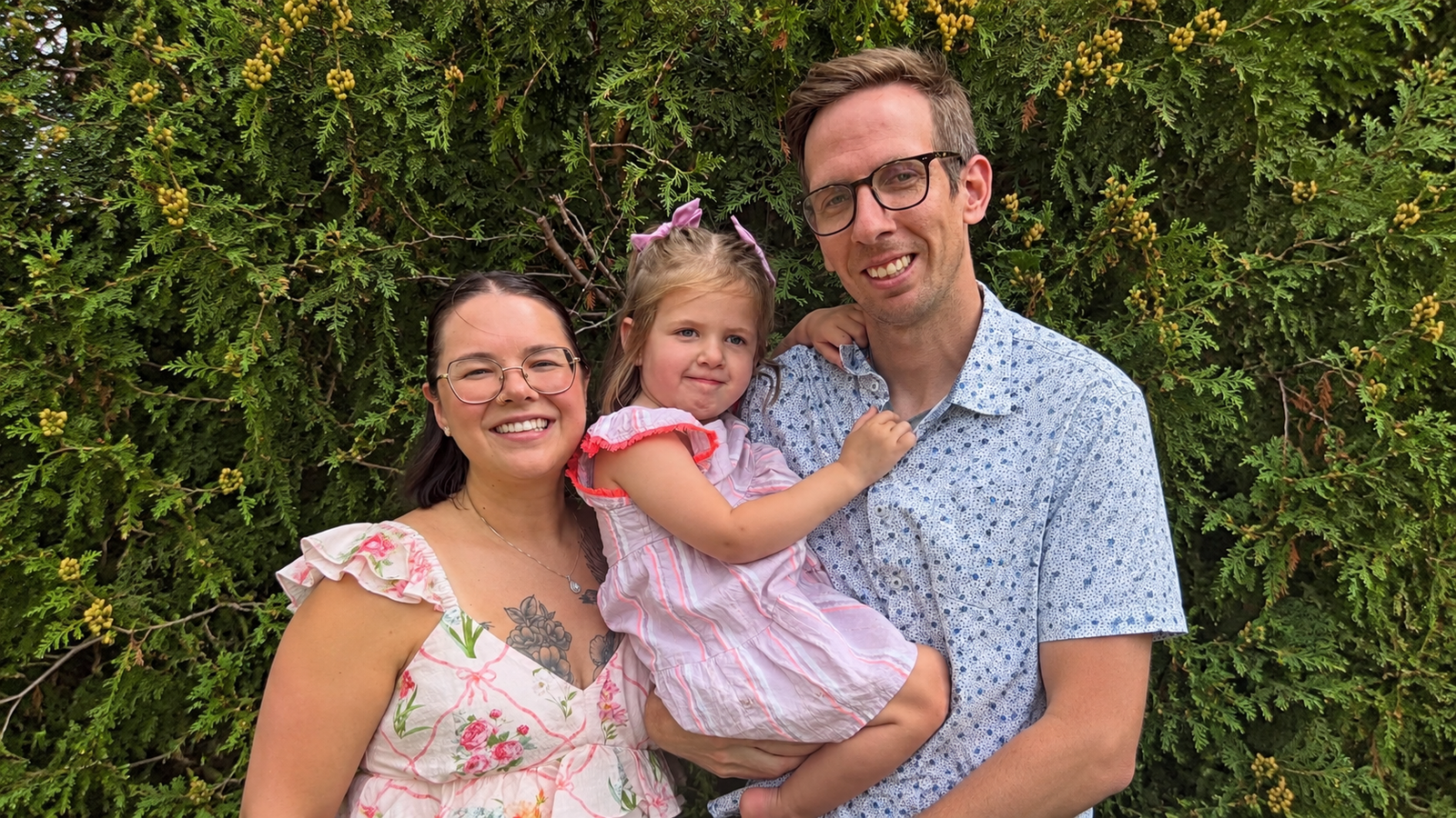 Rachael Lang smiles beside her husband, who holds their young daughter Delaney in a lavender dress and hair bows, in front of evergreen shrubs.
