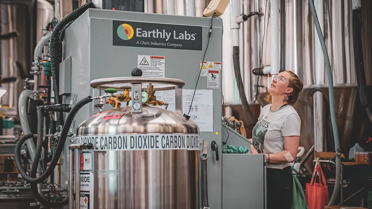 A brewer in safety glasses beside a stainless carbon-dioxide vessel and an Earthly Labs carbon-capture unit branded as a Chart Industries company, inside a brewery