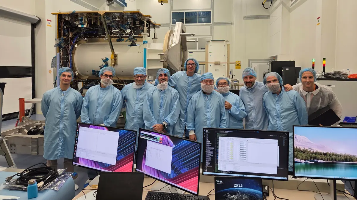 Ten engineers in blue cleanroom gowns and hairnets stand in a line, smiling in front of the gold-wrapped Ramses Core Module, with an overhead gantry crane above them and monitor screens showing test data in the foreground
