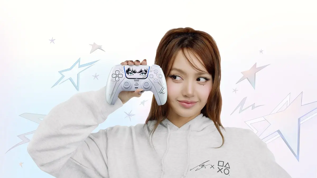 Lisa, in a pale grey sweatshirt, holding a limited-edition DualSense controller whose touchpad is printed with her eyes and fringe, against a pale backdrop scattered with stars.